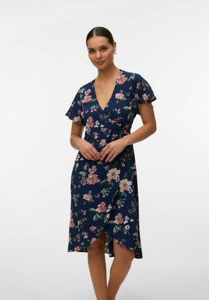 Kobieta o ciemnych włosach ubrana w granatową sukienkę w kwiaty z krótkimi, zwiewnymi rękawami, stojąca na jednolitym białym tle.