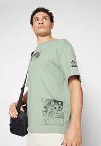 Ανοιχτό πράσινο βαμβακερό μπλουζάκι με γραφικά σχέδια στο μπροστινό και στο πλάι, χαλαρή γραμμή, κοντά μανίκια και λογότυπο "Star Wars" στο μανίκι.