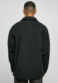Jachetă din fleece neagră, cu un material moale și texturat, croiala supradimensionată, guler rotund și umeri lăsați, fără hardware vizibil.