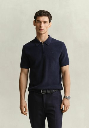 Junger Mann mit dunklem Kurzarm-Poloshirt, schwarzem Gürtel, dunkler Hose und silberner Armbanduhr steht vor einem hellen, schlichten Hintergrund.