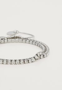 Sølv tennisarmbånd med klare firkantede edelsten satt i en dobbel rekke på en hvit bakgrunn, med en justerbar lås.