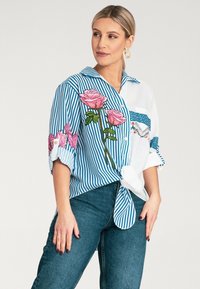 Pasiasta niebiesko-biała bluzka z kwiatowym haftem w odcieniach różu i zieleni. Posiada zapięcie na guziki z przodu oraz wiązanie w talii.