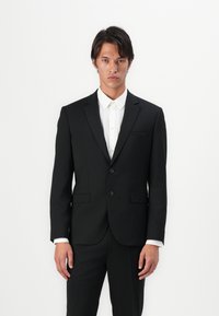 Costume noir à deux boutons avec une coupe ajustée, revers en pointe et poches avant. Chemise blanche en dessous, avec un col classique et fermeture à boutons.