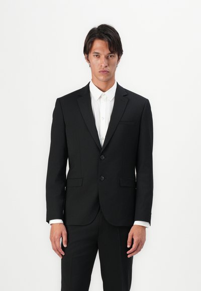 Costume noir à deux boutons avec une coupe ajustée, revers en pointe et poches avant. Chemise blanche en dessous, avec un col classique et fermeture à boutons.