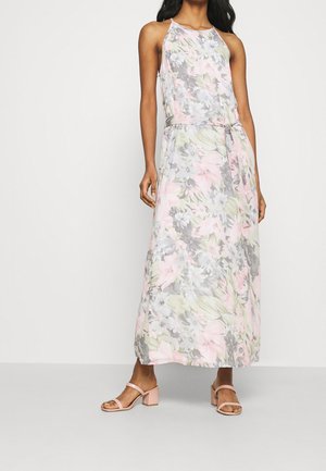 Vrouw draagt een mouwloze maxi-jurk met pastelkleurige bloemen en lichtroze sandalen met hak, staand tegen een effen witte achtergrond.