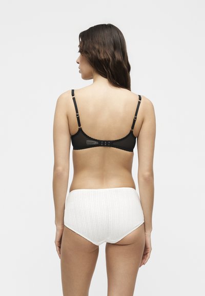 Hosszú barna hajú nő fekete melltartóban és fehér, magas derekú kötött fehérneműben áll, háttal nekünk, egyszínű háttér előtt.