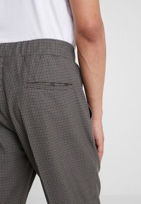 Gros plan sur une personne portant un pantalon à carreaux gris et marron avec une poche arrière, associé à une chemise blanche rentrée.