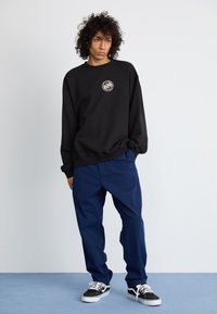 Sudadera negra de gran tamaño con un logo circular, combinada con pantalones azul marino y zapatillas negras, con cordones blancos y suela plana.