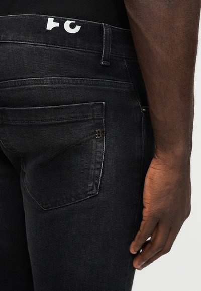 Gros plan sur la main droite d'une personne, détendue à côté de son jean en denim gris foncé, avec une poche arrière visible et une ceinture noire.