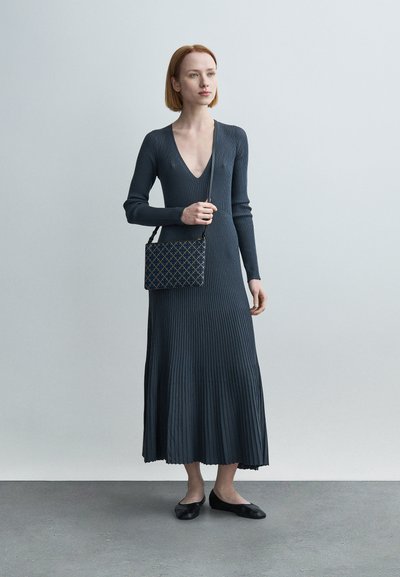 Robe longue côtelée gris foncé avec un décolleté en V profond et des manches longues. Associée à un petit sac à main à motifs et des ballerines noires. Texture lisse.