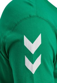 Grön bomulls-t-shirt med en mjuk textur, med tre vita nedåtriktade chevroner på vänster ärm. Reflekterar en sportig design.