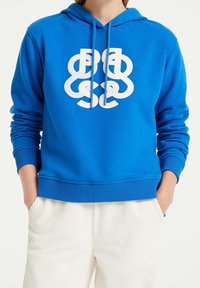 Blue hoodie with a front white graphic logo, drawstring hood, ribbed cuffs, and hem, made from soft cotton material. Paired with cream corduroy pants.