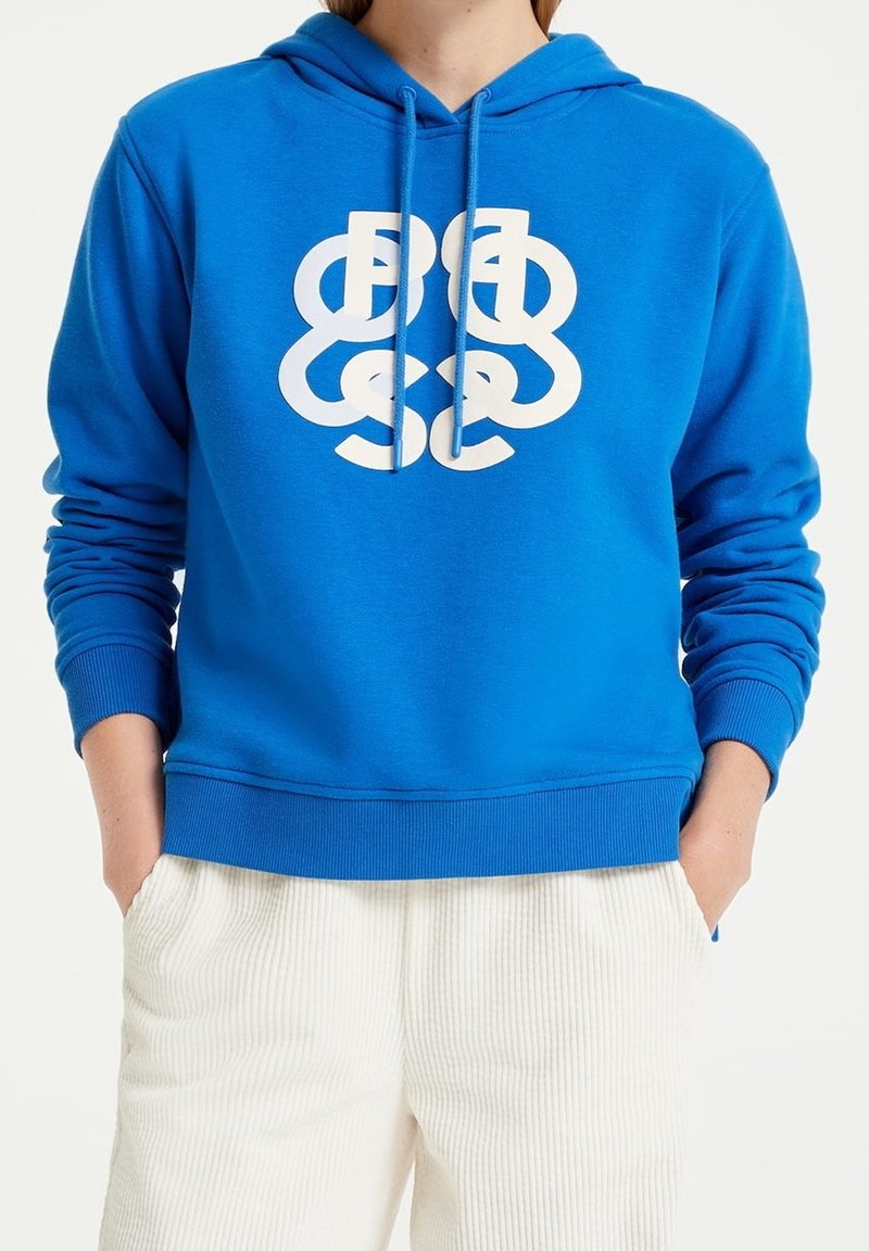 Blue hoodie with a front white graphic logo, drawstring hood, ribbed cuffs, and hem, made from soft cotton material. Paired with cream corduroy pants.