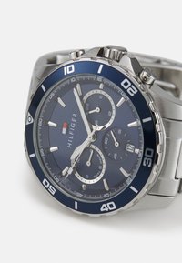 Montre chronographe en argent avec un cadran bleu marine, aiguilles lumineuses et trois sous-cadrans. Comprend un boîtier et un bracelet en acier inoxydable.