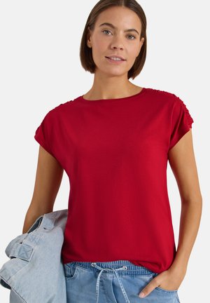 Jeune femme portant un haut rouge à manches courtes et un pantalon en jean bleu, tenant une veste en jean bleu clair dans sa main gauche.