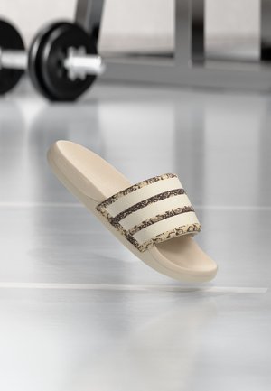 Sandale beige à enfiler avec trois bandes sombres à motifs sur la bride, posée sur un sol de salle de sport réfléchissant avec des haltères flous en arrière-plan.