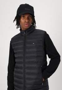 Black puffer vest with horizontal quilted design and stand-up collar, worn over a black long-sleeve shirt. Cap with "BOSS" logo in black.