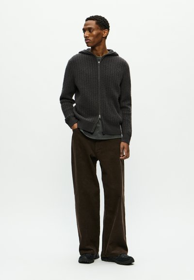 Sudadera de punto de color gris oscuro con cremallera y un patrón texturizado, combinada con pantalones de pana anchos de color marrón y zapatillas negras.