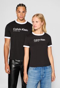 Juodos medvilninės marškinėliai su balta apykakle, kuriuose priekyje yra baltu spausdintu "Calvin Klein endless city" logotipu.