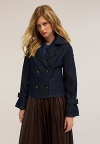 Abrigo peacoat de mezclilla oscura con diseño de doble botonadura, grandes botones y solapas anchas. Presenta adornos decorativos con lazo en las mangas.