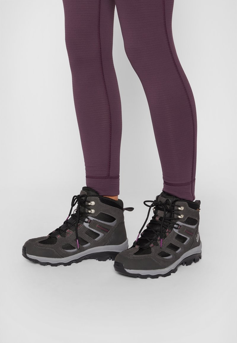 Jambes portant un legging violet et des chaussures de randonnée gris foncé avec des lacets noirs, des œillets en métal et des semelles robustes sur un sol gris clair.
