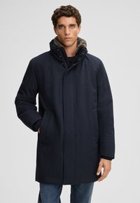 Cappotto blu navy con finitura testurizzata, cappuccio e tasche frontali. Presenta una chiusura a bottoni e un rivestimento interno in contrasto. Design elegante e su misura.