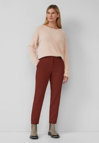 Vrouw staat met haar hand in de zak, gekleed in een lichtroze fluffy trui, roestkleurige slim fit broek en grijze enkellaarzen voor een effen grijze muur.