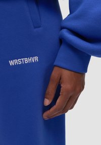 Niebieska bluza z teksturowanej tkaniny, z białym haftowanym logo "WRSTBHVR" nad kieszenią. Ręka dotyka materiału.