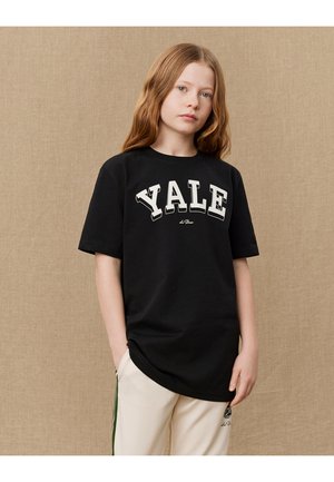 Svart bomull T-skjorte med stort hvitt "YALE" trykk, crew neck og avslappet passform, iført med kremfargede bukser med grønne sidestreker.