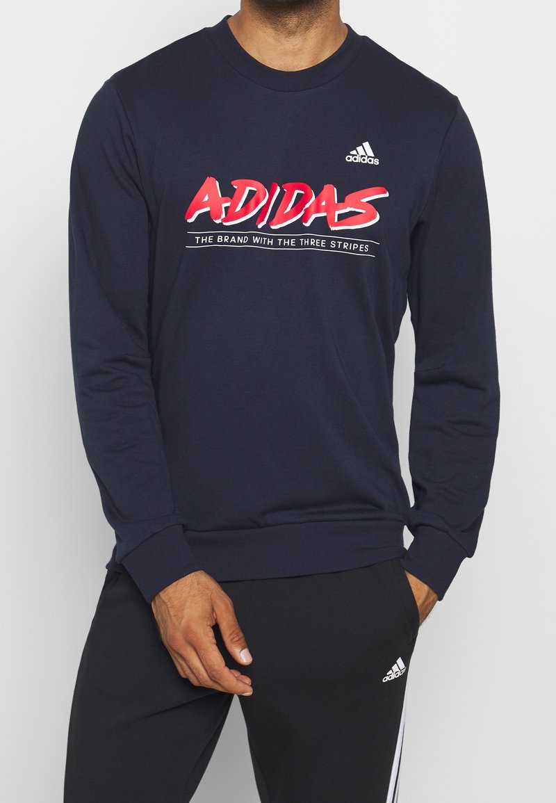 Homme portant un sweat Adidas bleu marine avec un logo rouge et un pantalon de survêtement noir Adidas avec des bandes blanches, debout avec une main dans la poche.