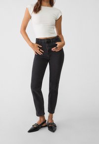 Mujer vestida con un crop top blanco ajustado, jeans negros de cintura alta y zapatos planos negros con hebillas, de pie con las manos en las caderas.