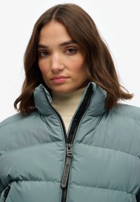 Lyseblå oppustelig jakke med høj krave og lynlås. Har synlige sømme og et logo på lynlåsen. Bæret over en beige sweater.