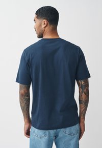 Tamsiai mėlynas medvilninis marškinėlis su apvalia iškirpte ir trumpomis rankovėmis, matomas iš nugaros, su tiesia apatine siūle ir be matomų logotipų.