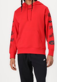 Röd sweatshirt med huva, prydd med svarta logotryck på ärmarna, en framficka och ribbade muddar. Matchad med mörka byxor.