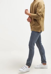 Chemise beige à manches longues, jean gris slim fit, et baskets blanches. Le jean présente une coupe fuselée, avec un look épuré et simple.