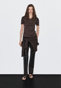 Top marrone a maniche corte abbinato a un avvolgente in maglia e pantaloni in pelle neri a gamba dritta. Sandali neri piatti completano l'outfit. Sfondo neutro.