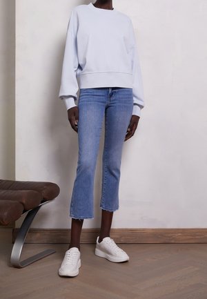 Personne portant un sweat-shirt blanc oversize, un jean bleu clair raccourci avec des ourlets effilochés, et des baskets blanches, debout à l'intérieur.