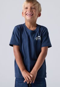 Marineblå kortermet T-skjorte laget av bomull, med et grafisk motiv av kryssete rutete flagg på brystet. Enkel rund halsdesign.