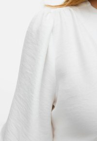 Top bianco in tessuto testurizzato con una vestibilità rilassata. La manica presenta un leggero dettaglio a sbuffo, che sottolinea la sua struttura morbida.