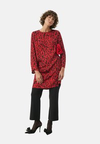 Rød leopardmønstret tunika med lange ærmer, parret med sorte cropped bukser. Har et tekstureret rødt tilbehør og høje sorte sko.