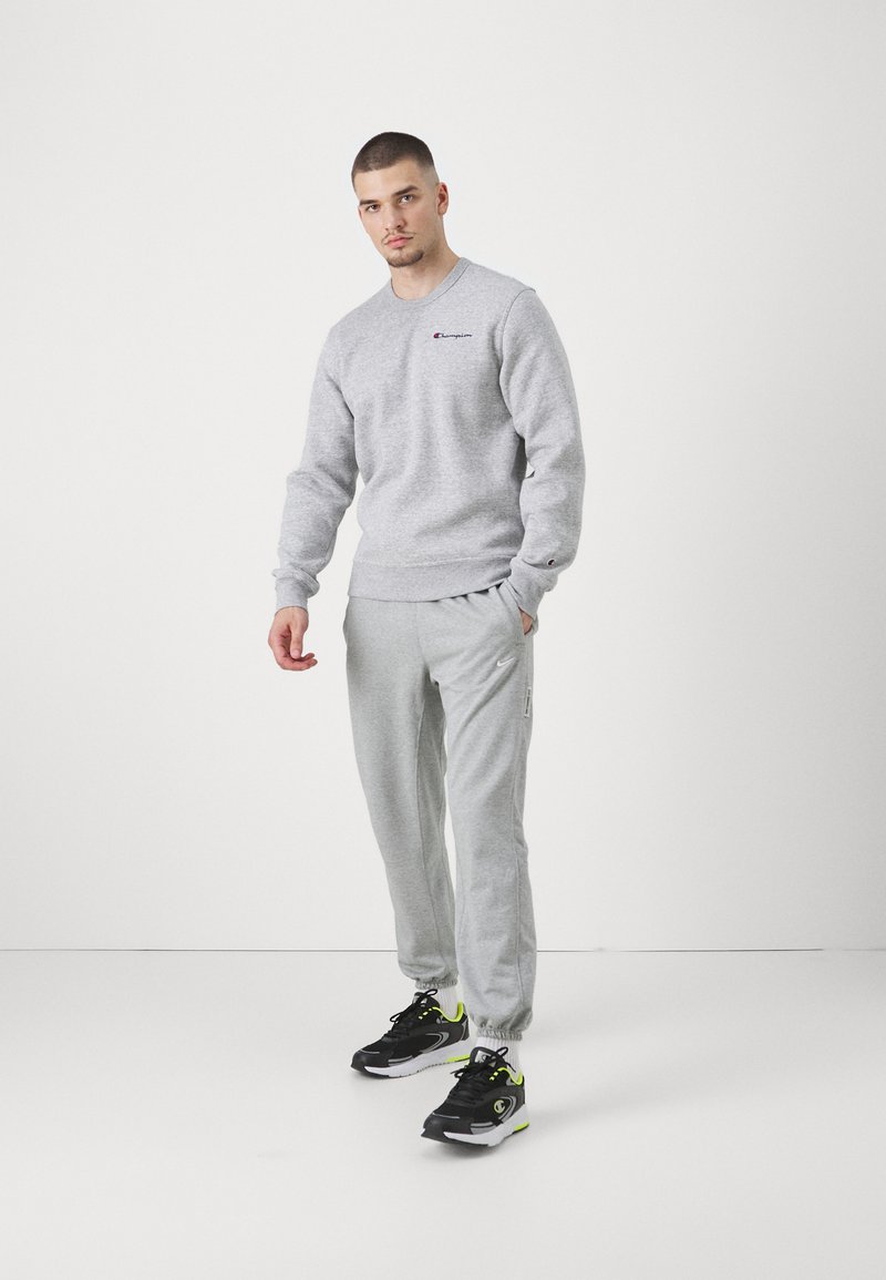 Sudadera gris con cuello redondo y logotipo, combinada con pantalones de chándal grises con bolsillos laterales. Zapatos negros con acentos en neón.