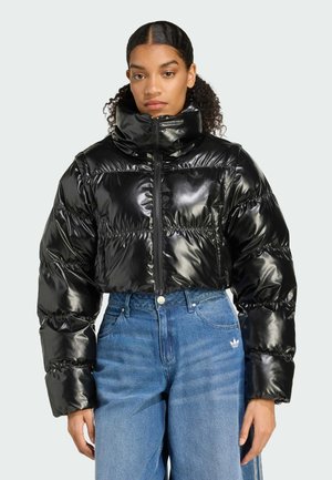 En kvinna med lockigt hår bär en kort, glänsande svart pufferjacka och blå jeans, stående mot en ljus bakgrund.