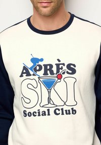 Felpa girocollo color crema con maniche blu navy. Stampa grafica con uno sciatore, un cocktail e il testo "Après Ski Social Club" in colori vivaci.