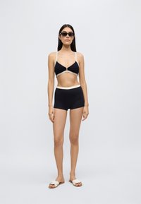 Svart todelt badedrakt med beige detaljer, med en trekantet topp med tynne stropper og en bikinitruse med høyt liv. Modellen har på seg solbriller og sandaler.