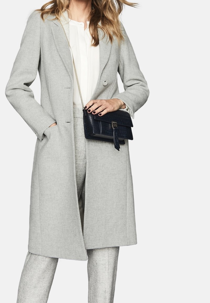 Světlý šedý saka s kapsami, kombinovaný s bílou halenkou a texturovanými světlými šedými kalhotami. Drží černou kabelku s krokodýlím vzorem.