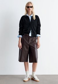 Schwarze Strickjacke über einem hellblauen Hemd, braune Leder-Shorts, weiße Socken und beiges Sneakers. Die Accessoires umfassen dunkle Sonnenbrillen.