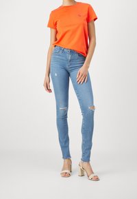 Camiseta de algodón naranja de manga corta con un pequeño logo, combinada con jeans skinny de color azul claro que presentan desgastes en las rodillas y un dobladillo deshilachado.