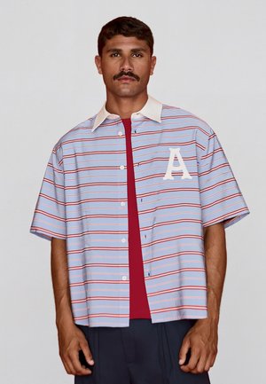 Bajszos férfi világoskék rövid ujjú csíkos ingben, fehér gallérral és nagy fehér "A" betűvel a mellkasán, piros aláöltözet fölött.