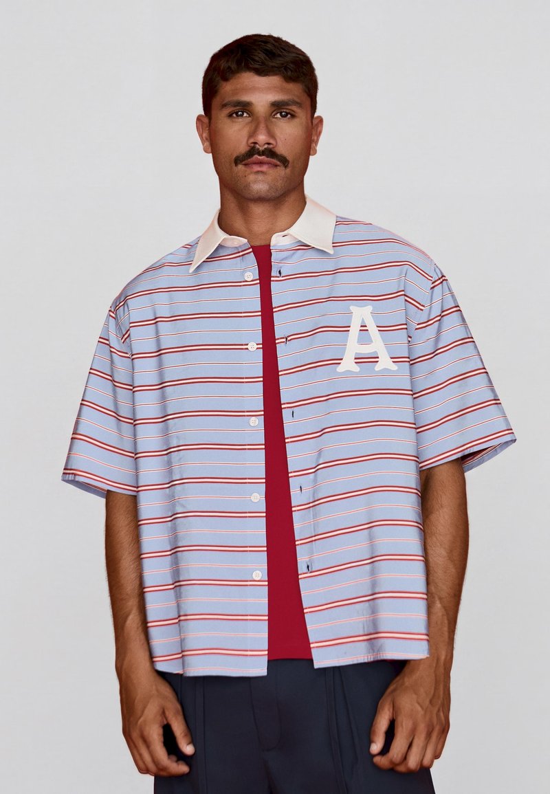 Homme avec moustache portant une chemise rayée bleu clair à manches courtes avec col blanc et un grand « A » blanc sur la poitrine, par-dessus un sous-vêtement rouge.