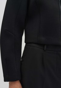 Veste noire avec un design structuré, un tissu texturé et un col haut. Associée à un pantalon noir taille haute.